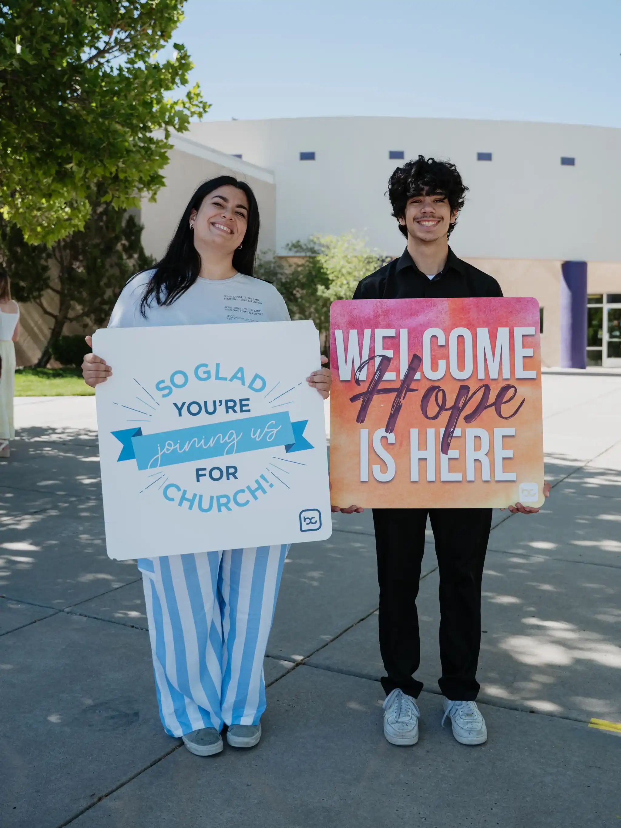 Two host team members smiling with welcome signs outside Believer Church