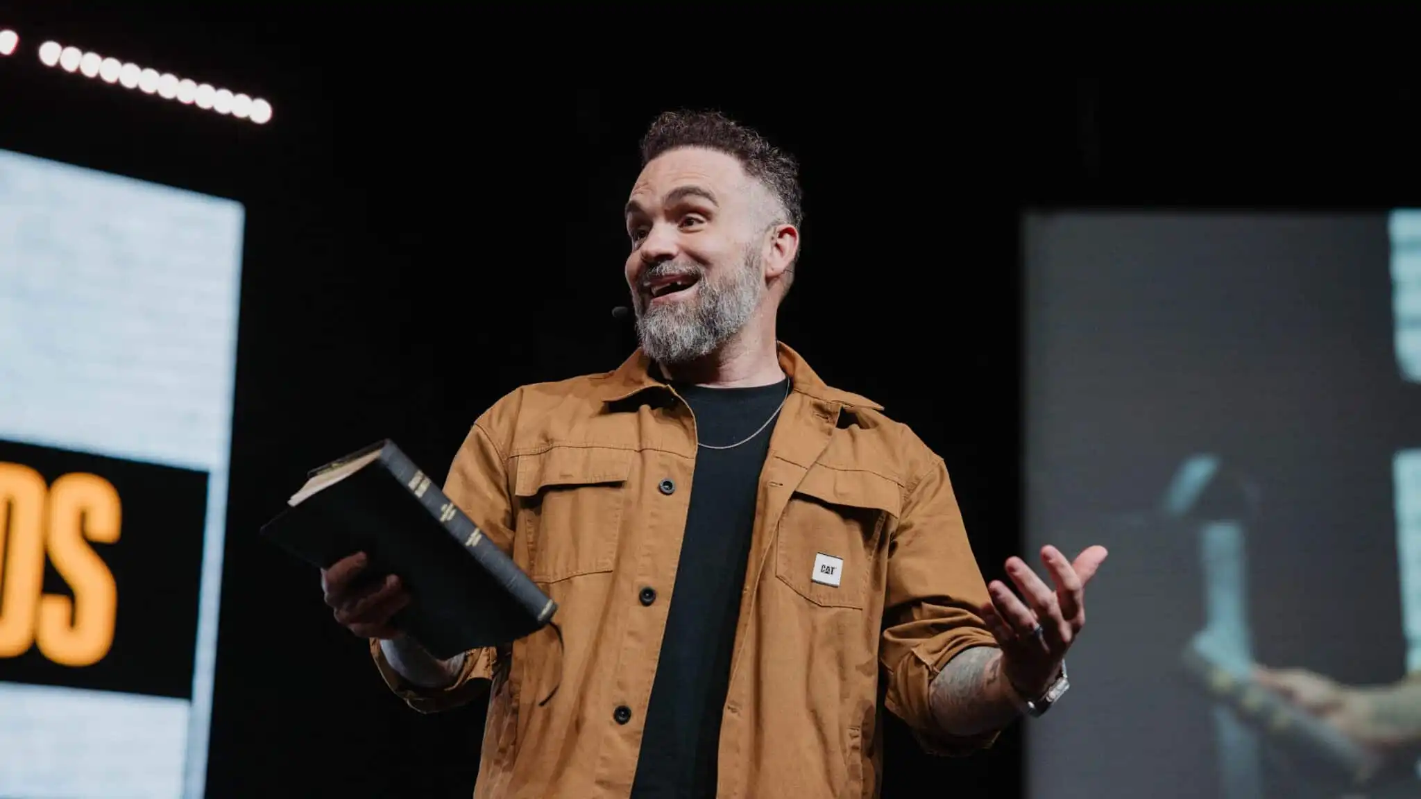 Pastor smiling and gesturing while holding a Bible on stage